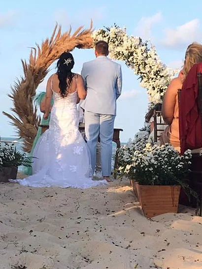 Noiva de costas diante do arco de flores na areia, com o mar ao fundo